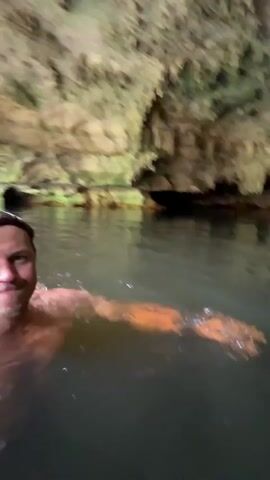 A person with short blonde hair is swimming in a body of water inside a cave. The cave walls are visible above and behind the person, with stalactites hanging from the ceiling. The water is dark and murky. The person is looking up at the cave ceiling with an astonished expression.