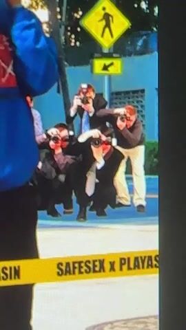 The video shows a group of photographers, possibly paparazzi, crouching and taking pictures. They are wearing suits and ties, and some have cameras with flashes. In the background, there is a yellow pedestrian crossing sign and a street. A yellow caution tape with the words "SAFESEX x PLAYAS" is visible in the foreground.
