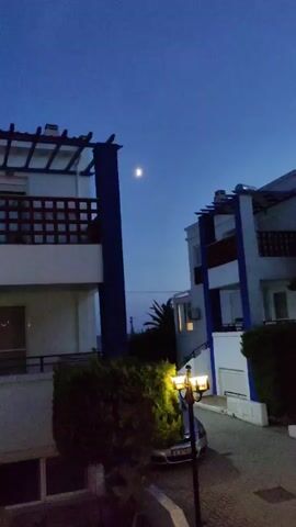 The video shows a twilight scene with a crescent moon visible in the deep blue sky. Two modern, white buildings with blue accents are positioned in the foreground, partially obscuring a driveway. A streetlamp with two illuminated bulbs stands in the middle of the driveway, casting a warm glow. A dark-colored car is parked in the driveway, partially visible behind the streetlamp. Lush green bushes and trees are planted around the buildings and in front of them. The overall atmosphere is peaceful and serene, suggesting a quiet evening.
