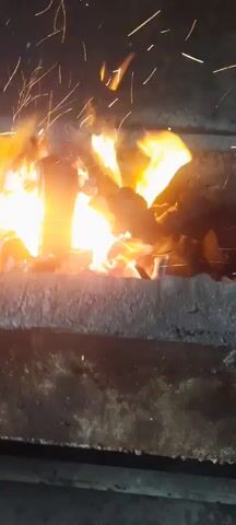 A close-up shot shows a wood-burning fire in what appears to be an outdoor oven or fireplace. The flames are bright orange and yellow, with sparks flying upward. The fire is contained within a dark, stone-like structure. In the foreground, a person's hand, adorned with a thin bracelet, reaches into the frame, seemingly to adjust the logs or embers. The overall environment is dark, suggesting it is nighttime or a dimly lit area. The video captures the dynamic nature of the fire and the interaction with it.