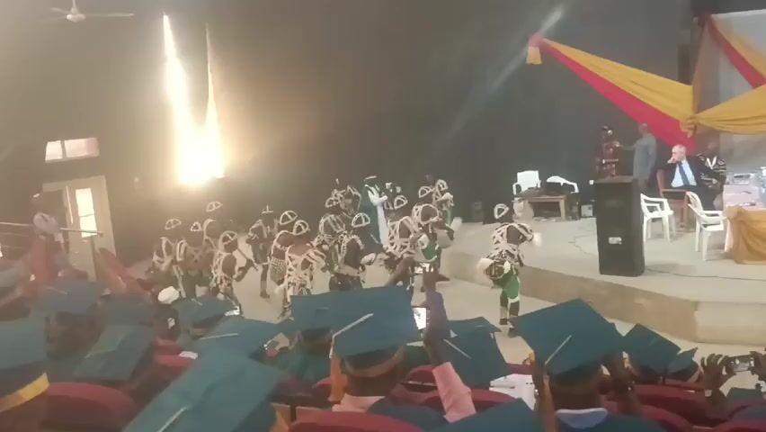 A video shows a group of people dressed in traditional African dance attire performing in front of an audience. The dancers are wearing white and black patterned outfits, headgear, and some have green accents. They are moving in unison, with some holding instruments. The audience is primarily composed of people in graduation gowns and mortarboards, many of whom are holding up their phones to record the performance. The room appears to be a large hall with a stage at the front. There are individuals seated on the stage and draped fabric of red and yellow hues.  The lighting in the hall is somewhat dim.