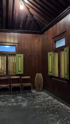 This media file appears to be a short video or series of still images showcasing the interior of a traditional Javanese house in Banguntapan, Indonesia. The setting is characterized by rich, dark wood paneling on the walls and ceiling, creating a warm and somewhat somber atmosphere.

The scene features several distinct objects and architectural elements. In the foreground, the floor is a dark, textured concrete. Towards the left, there's a tall, ornate green cabinet filled with various decorative items. In front of a set of wooden shutters, three simple wooden chairs are arranged. A tall, woven cylindrical object, possibly a basket or decorative vase, stands in the corner.

Further into the house, a large wooden pillar is prominent. Through an open doorway, another room is visible, also adorned with green and yellow accents on cabinets and doors. The overall decor suggests a historical or cultural exhibit, possibly a museum or a well-preserved heritage home.

Notable details include the traditional Javanese architectural style, with intricately designed wooden elements and the use of vibrant green and yellow paint as accents on some furniture and fixtures. The lighting is somewhat dim, suggesting it might be late afternoon, evening, or simply an indoor space with limited natural light. No people are visible in the frame. There are a few small signs with text, but the text is too small and indistinct to be read.