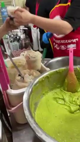 The video shows a busy food stall in what appears to be a market or food court setting. The stall is selling what seems to be desserts or snacks. A woman wearing a red apron with writing on it is working at the counter, wearing blue gloves, scooping something out of a bowl with a pink-handled utensil. There are various ingredients and containers visible on the counter, including a large bowl of a bright green mixture, several smaller containers filled with different toppings or ingredients (including what looks like jellies or fruits), and rows of pre-made snacks or desserts in cups displayed in a refrigerated case. Other people are visible in the background, either working at the stall or as customers. A QR code payment option is visible on the counter. The environment is bustling with activity and appears to be a popular spot for food.