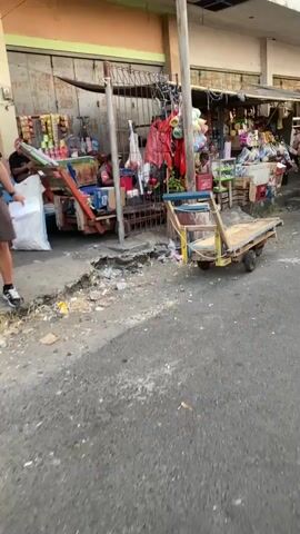 The video shows a street scene in front of a row of small shops. On the left, a person wearing a dark shirt and shorts stands near a cart filled with goods, holding a white piece of paper. To the right of this, there is a cart with a blue seat and a wooden platform. The ground in front of the shops is littered with trash, and there is a large crack in the pavement. The shops themselves are filled with various items, including packaged food, drinks, and household goods. The overall impression is of a busy, perhaps somewhat disorganized, marketplace.