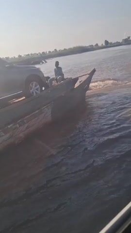 Here's a description of the video:

The video shows a car being transported on a makeshift raft or boat across a body of water. The raft appears to be constructed from wooden planks or logs, and the car is positioned on it. There are two or three people on the raft with the vehicle. They appear to be guiding or overseeing the transportation. The water has a brownish tint. The environment seems rural. The sky is bright, suggesting it is daytime. The raft is creating ripples in the water as it moves.