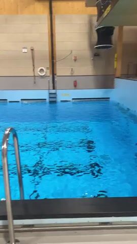 An indoor training facility in Horten, Norway, is shown featuring a large swimming pool filled with clear blue water, with lane markers visible on the pool bottom. The room has high ceilings with exposed wooden beams and light-colored concrete walls.

In the foreground, metal pool ladders or railings are visible. The primary activity involves a large, orange, inflatable, enclosed liferaft or survival capsule, which is initially suspended from a ceiling hoist. As the video progresses, the liferaft is slowly lowered and then released, dropping into the water with a significant splash.

Several individuals, dressed in bright yellow survival suits, are already present in the pool. Some are seen floating in smaller yellow and orange inflatable rafts. They approach the deployed liferaft, and towards the end of the clip, some trainees are shown climbing into the orange capsule.

The scene depicts a sea survival or emergency response training exercise. Notable details include a clock and the letters "RSE (+)" prominently displayed on the far wall, possibly indicating the facility's name. Yellow warning signs are placed along the pool's edge, one clearly visible stating "STUPING FORBUDT" (No Diving). The facility is brightly lit, suggesting daytime conditions.