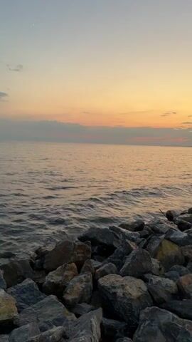 The video shows a rocky shoreline with the ocean in the background during sunset. The water is calm with gentle waves lapping against the rocks. The sky is painted with soft hues of orange, pink, and yellow, indicating the sun is setting or has recently set. A thin strip of clouds is visible on the horizon. In the distance, a faint structure, possibly a pier or a building, can be seen on the far right side of the frame. The foreground is dominated by large, grey and brown rocks, forming a natural barrier along the coast. The overall ambiance is peaceful and serene.