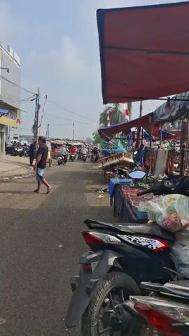 A street market is visible with stalls on the right side and a street on the left. A person is walking on the left side of the street. Several motorbikes are parked on the right side of the street, in front of the market stalls. The stalls have red awnings, and some appear to be selling food or goods. There are also more people and motorbikes further down the street, suggesting a busy market environment. The sky is clear and the sun is shining.
