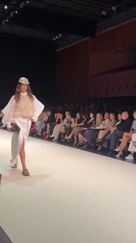 A fashion show is in progress. A female model walks down a white runway wearing a white skirt, a sheer white shirt with voluminous sleeves, and a sparkling sequined vest. She also wears a light-colored cap and carries a dark clutch bag. She is wearing light-colored sandals. Spectators are seated on both sides of the runway, observing the model. The environment appears to be an indoor auditorium with dark walls and stage lighting.
