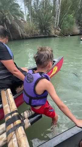 A young boy wearing a purple life vest and red swim trunks is seated on the edge of a red canoe, with his feet in the green water. His left leg is extended, and his right leg is bent. He is facing away from the camera, looking towards the right. To his left, a person wearing a blue and black life vest is also in the canoe, with their back partially to the camera. In the foreground, a raft made of logs is visible. The canoe is floating in a river with lush green vegetation and trees along the bank. The water appears calm.