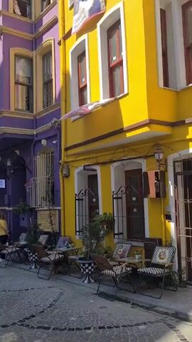 The video captures a vibrant street scene in Istanbul, Turkey, characterized by brightly colored buildings and outdoor seating for cafes. Dominating the frame are two buildings, one painted a striking purple and the other a cheerful yellow, their facades adorned with traditional bay windows. In front of these buildings, several small tables and chairs are arranged, some with black and white checkered cushions, suggesting a bustling cafe environment.

The time of day appears to be daytime, with clear skies and bright sunlight casting shadows on the cobblestone street. The weather seems pleasant and warm, encouraging outdoor activity. Several people are visible, some seated at the cafe tables, while others walk along the street. One woman in the foreground, wearing a white t-shirt and beige shorts, strikes a pose for the camera, her expression suggesting a lighthearted and enjoyable moment. Another woman in a yellow t-shirt walks by the cafe, and other individuals are seen in the background, contributing to the sense of a lively urban setting.

A sign on the purple building reads "NOSTALGIC" in white lettering. Another sign, partially visible on the yellow building, displays text in what appears to be Turkish, possibly indicating a business name or advertisement. The overall atmosphere is one of relaxed tourism, with the colorful architecture and the presence of people enjoying the outdoors contributing to a cheerful and inviting ambiance. The juxtaposition of the vividly painted buildings and the everyday activity of a street cafe creates a picturesque urban landscape.