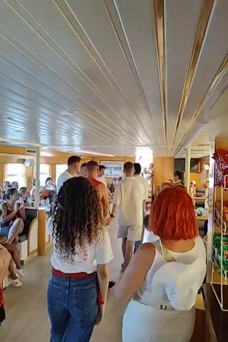 The video shows the interior of a boat, likely a passenger ferry or cruise ship, with a group of people. The camera is positioned at the rear of the boat, looking forward. The ceiling is paneled with white material and decorated with gold trim. The walls are also white, with windows visible on the left side.

In the foreground, two women are facing away from the camera. One has long, curly brown hair and wears a white t-shirt with red lettering and blue jeans. The other has short, bright orange hair and wears a white top with a matching hat.

Further forward, a group of men in casual clothing, including t-shirts and shorts, are walking away from the camera. Other passengers are seated on benches along the sides of the boat, looking towards the front. Some people are standing and talking. The overall atmosphere appears to be relaxed and social. The lighting is bright, suggesting it is daytime.
