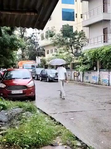 A person walks across a wet street, holding an umbrella. The person is wearing a white shirt and grey pants. A red car is parked on the left side of the street. Other cars are also visible on the street. Buildings and trees are in the background. The scene appears to be during a rainy day.