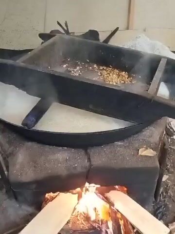 A video shows a traditional cooking setup where food is being prepared over an open fire. A large, dark, rectangular metal pan is suspended over a brick stove where burning wood provides heat. The pan appears to be divided into two sections. In the right section, there are small, golden-brown kernels, possibly corn or nuts, being heated. In the left section, a white, frothy liquid, likely boiling or simmering, occupies the majority of the pan. Steam rises from both sections of the pan, indicating the cooking process is underway. The surrounding environment appears to be rustic, with a dirt or concrete floor and possibly wooden supports or structures visible in the background. The focus is on the cooking activity itself.