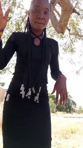 A young woman is seen in a short outdoor video clip. She is standing under a tree, dressed in a black, long-sleeved top and a black pencil skirt. She has braids with silver-colored beads woven into them. The sun shines brightly behind the tree, creating a somewhat high-key shot. In the background, there is a building, some grass, and another tree. The woman adjusts her braids, turns slightly to the side, and then smiles at the camera while making small dance-like movements with her arms.