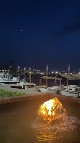 The video opens with a close-up of a fountain in a shallow pool of water. The water is dark, and a bright, fiery object is visible in the center, with water splashing around it. The camera then pans left to reveal a harbor at dusk or night. Several boats are docked along a walkway. The sky is dark blue with some clouds. In the background, lights from buildings and more boats are visible along the shoreline. As the camera continues to pan, a person in a red shirt is seen sitting on the dock, and another person in a white dress is standing further down the walkway. The camera then pans slightly to the right, showing more boats and palm trees along the waterfront. The scene appears to be a marina or harbor area at twilight or nighttime.