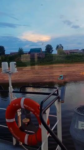 The video shows a view from a boat on a river. In the foreground, a life preserver is visible, attached to the railing of the boat. The life preserver is orange with white stripes and has Russian text on it. Behind the life preserver, there is a white structure with lights on top, likely part of the boat's navigation system. The water of the river is dark and calm. In the background, across the river, is a sandy bank lined with vegetation. Further inland, there is a small village with wooden houses and green roofs. The sky is partly cloudy with hints of sunset colors in the distance. The overall impression is a peaceful, rural scene on a river at dusk.