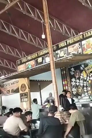 A group of people are gathered inside what appears to be a restaurant or food stall. In the background, a sign reads "SMEA PREMIUM TAKENGO" with several framed images below it. A woman wearing a hijab and an apron is visible behind a counter, appearing to be preparing food or drinks. To her right, two men in dark jackets are standing. In the foreground, several people are seated at tables, facing the counter. One person is holding a smartphone. The environment is covered with a corrugated metal roof supported by metal beams and a large wooden pole in the center.