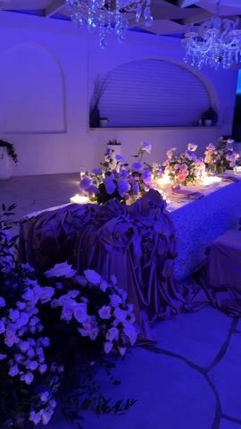 The scene depicts a formal dining setup under blue lighting. The central focus is a long, low table covered in a lace tablecloth and draped with a satiny, crumpled fabric. Several floral arrangements, primarily of white and pink roses, are placed along the length of the table, interspersed with lit candles. Two crystal chandeliers hang from the ceiling above the table. The chairs are low, cushioned, and also covered in the same satiny fabric. The room has a Mediterranean architectural style, with white walls and arched alcoves. The floor is made of irregular stone slabs. The overall atmosphere is luxurious and romantic.