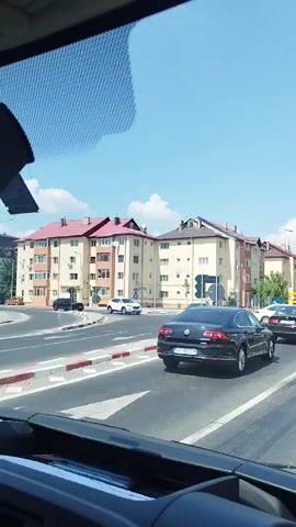 The video shows a street scene with several cars and buildings under a clear blue sky. In the foreground, the dashboard of a vehicle is visible. In the middle ground, a black sedan is driving on the road, followed by a truck with a white cage and green branding on its side. The truck is carrying various items, including what appears to be a spare tire. Across the street, several apartment buildings with red tiled roofs are visible. Traffic signs and lights are also present. The camera appears to be mounted inside a moving vehicle, capturing the view through the windshield.