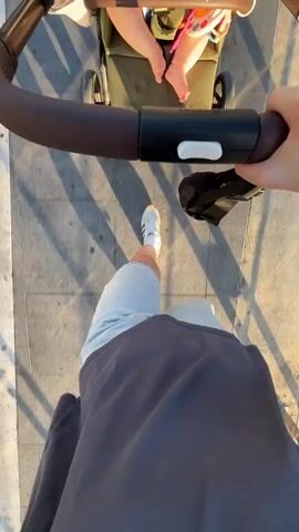 The video is a first-person perspective of someone pushing a stroller with a baby in it. The person pushing the stroller is wearing a dark sweatshirt and light-colored shorts, with white sneakers. The baby is visible in the stroller, wearing a pink and white outfit and has small feet. The stroller appears to be a dark color. They are walking on a paved surface, likely a sidewalk, with a shadow cast on the ground. The lighting suggests it is either early morning or late afternoon, with the sun casting long shadows. The background is out of focus, but appears to be outdoors, possibly a park or promenade. The camera is angled downwards, focusing on the stroller handle and the legs of the person pushing it.