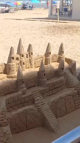 The video begins with a close-up, high-angle shot of an elaborate sandcastle on a sandy beach. The sandcastle has multiple towers, walls, and intricate details resembling doorways and windows. In the background, the ocean is visible with waves, and several beach umbrellas and people are scattered across the sand.

The camera then pans slightly to the left, showing more of the beach area, including the ocean and more beachgoers. A man, shirtless and with a tattoo on his left shoulder blade, walks into the frame from the right and moves down a sandy incline towards the sandcastle. He appears to be looking at the sandcastle and potentially interacting with it, although his actions are not clearly visible from this angle. The overall environment is a bright, sunny day at the beach.