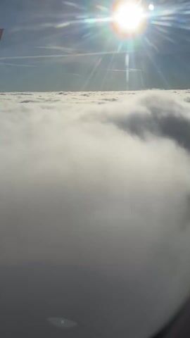 The video shows a view from an airplane window. The sun is shining brightly in the sky, above a thick layer of clouds. Part of the airplane's wing is visible in the upper left corner of the frame. The clouds are dense and white, creating a blanket effect. The video seems to be taken during the daytime. There is an announcement being made over the aircraft's speakers.