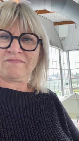 A woman with short blonde hair and black glasses stands in a large, bright indoor space. She wears a black sweater. Behind her is a tiled pool, surrounded by lounge chairs, and umbrellas. Large windows in the background show trees and distant buildings. The ceiling is high, with exposed wooden beams and ventilation ducts. The woman looks around, capturing the scene with the camera.