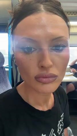 A woman is sitting on a train. She is fair-skinned, with dark hair pulled up into a bun. She is wearing a black t-shirt, eye liner, lip gloss, and earrings. She is looking toward the camera, turning her head slightly to the side, and speaking. An earbud is visible in her left ear. Other people are visible in the background.