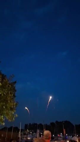 A fireworks display is taking place against a dark blue sky. Several fireworks have exploded, creating bursts of light and sparks. In the foreground, the backs of several people's heads are visible as they watch the display. The scene appears to be taking place near a body of water, with boats and a treeline visible in the background. The overall atmosphere is festive, indicating a celebration or event.