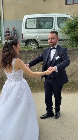 A bride and groom are dancing in an outdoor scene. The bride is wearing a white wedding dress with intricate floral designs, and her hair is styled with a floral headpiece. The groom is in a navy blue suit with a black bow tie. They are holding hands and dancing with some twirling motions. There's a concrete wall and a chain-link fence behind them, and a white van with an image on it is parked nearby. Two figures are partially visible behind the fence. The ground appears to be a paved surface, possibly a road or driveway. The overall lighting suggests it is daytime.