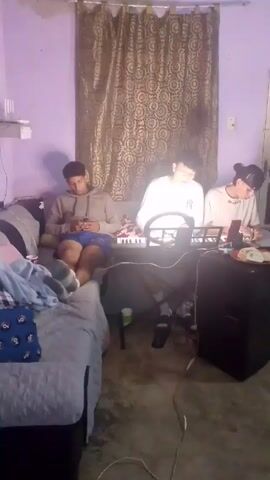 Three young men are in a room with lilac walls. One sits on a bed on the left, looking at his phone.  The bed is covered with a blue and gray patterned blanket.  Another young man is seated in front of a keyboard playing music. The third young man is seated to the right of the keyboard, appearing to be eating or writing while looking at a phone.  Behind the men is a brown and gold patterned curtain.  The room is cluttered with miscellaneous items.  A black speaker sits on the floor to the right.