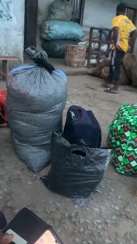 The video pans across a dusty outdoor area, possibly a marketplace or transport hub. In the foreground, several large bags are piled up, including a massive gray mesh bag filled with what appears to be produce, and a black plastic garbage bag. To the right of these are two suitcases, one dark blue and another with a plaid pattern. Further to the right, there is a brightly colored green bag with a red and white pattern, and a cardboard box. In the background, a person wearing a yellow t-shirt and dark shorts is walking away from the camera. Other large bags, some appearing to be burlap sacks, are visible in the background, along with a white van. A person in a blue and white checkered shirt is seated on a wooden bench to the left. The overall scene suggests preparation for transport or unloading of goods.