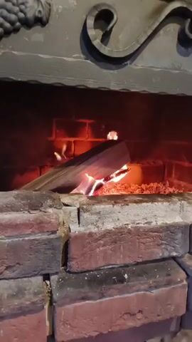 A close-up video shows a fire burning in a brick fireplace. The logs are burning brightly with orange and yellow flames and some glowing embers. The fireplace has a metal mantelpiece with decorative scrollwork. The camera is positioned at eye level, looking into the fireplace.
