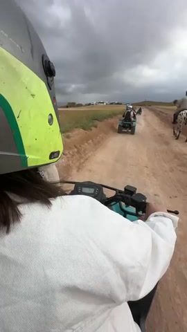 The video captures a first-person view from an ATV, traveling down a dirt road in a rural, open landscape. The helmet of the person filming, which has "LAZK" visible on its side, is prominently featured in the foreground. Ahead, another ATV can be seen on the road. To the right, a man rides a distinctively patterned horse, possibly an Appaloosa, with black spots on a white coat. The rider wears a helmet and turns to look towards the camera briefly. The horse is equipped with a traditional-looking saddle and bridle. The terrain is dry and dusty, flanked by fields of dry grass and distant hills or mounds under an overcast sky, suggesting cloudy weather during the daytime. The scene depicts a recreational outing involving both ATV and horseback riding on a dirt track, likely in the Lalla Takarkoust region of Morocco.