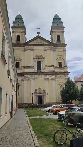The video shows a wide shot of a church with two bell towers, a tan facade, and a green roof. In front of the church, there is a paved walkway, a grassy area, and several parked cars. A bicycle is parked in the foreground. The scene appears to be in a European town, with cobblestone streets and traditional architecture. The weather is cloudy but bright. In the second half of the video, the camera pans to the right, showing more of the parked cars and people interacting with them, as well as a tall column with a statue on top in the distance. The video ends with a shot of a red car driving away on the cobblestone street.