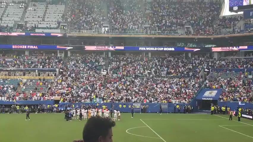 The video shows a wide shot of a soccer stadium filled with a large crowd of spectators. The field is visible in the foreground, with a group of players gathered in the center. The stands are packed with people wearing a variety of clothing. Large banners are displayed throughout the stadium, with one prominently showing "FIFA WORLD CUP 2026" and another indicating "BRONZE FINAL." The overall atmosphere appears to be one of a major sporting event.
