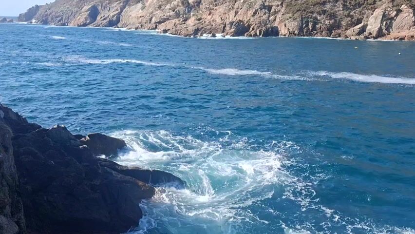 The video shows a coastal scene with blue water, rocky cliffs, and breaking waves. The water is deep blue with white foamy waves crashing against dark, jagged rocks in the foreground. In the distance, reddish-brown cliffs extend into the water, with some greenery visible on top. The water has some streaks of white foam further out, possibly indicating stronger currents or turbulence. The scene is bathed in sunlight, with a clear sky suggesting a sunny day.