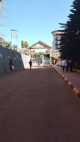 The video shows an asphalt road leading towards the entrance of Chuka University. The entrance has a gate and an archway with the university's name. People are walking along the road and through the gate. On the right side of the road, there is a small strip of green grass bordered by black and yellow concrete.  On the left, there is a tall corrugated metal wall covered with posters. The sky is blue and clear. A tall evergreen tree is visible on the right side of the frame.