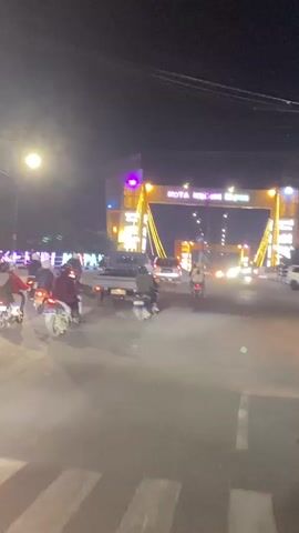 The video shows a nighttime street scene with a large, brightly lit archway that appears to be an entrance to a city or a significant landmark. The archway is adorned with yellow and purple lights. Several motorcycles and a small pickup truck are visible on the road, suggesting traffic flow. Some individuals are riding the motorcycles. The road surface is paved, and there are streetlights illuminating the area. The overall atmosphere is that of a busy urban environment at night, illuminated by artificial lighting. The text on the archway, partially obscured by lights, appears to read "KOTA KEDIRI" in large yellow letters, with additional text below.
