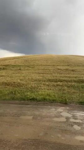 The video captures a panning shot of a rural landscape in Karagayl, Kazakhstan. The scene depicts a vast, rolling grassy hill under a dramatic sky. The weather appears to be changing, with dark, heavy clouds on the left side of the frame contrasting with brighter, sunlit patches breaking through on the right. This suggests a storm may be approaching or has recently passed.

In the foreground, a dirt road is visible, marked by numerous puddles of water, indicating recent rainfall. The road is uneven and appears to be a rural track rather than a paved surface. The hill itself is covered in dry, yellowish-brown grass, with some patches of greener vegetation closer to the road. The light suggests it might be late afternoon or early evening. The overall mood is one of natural beauty, with a touch of the dramatic due to the atmospheric conditions. There are no people or man-made objects other than the road itself. The expansive view emphasizes the open, undeveloped nature of the setting.