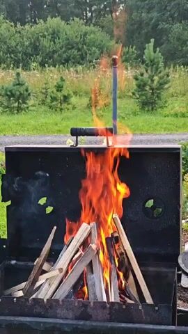 The video shows a campfire burning in a metal grill. The fire is made of wood and is producing a lot of flames. The grill is black and is placed in an outdoor setting with trees and grass in the background. There is a metal pole extending from the grill with a handle on top.