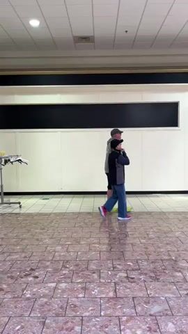Two individuals are walking side-by-side through a well-lit indoor shopping mall. The person on the left, wearing a grey hoodie, black shorts, a baseball cap, and lime green sneakers, is walking slightly ahead. The person on the right, wearing a dark vest over a grey long-sleeved shirt, blue jeans, and a black baseball cap with pink-soled sneakers, is walking slightly behind and to the left of the first individual, with their hands near their face. The floor is tiled with a pattern of pinkish-brown and cream-colored tiles. The walls of the mall are white, with a dark horizontal band across the upper portion. In the background, there are store fronts, tables and chairs, and other shoppers. The overall environment suggests a commercial indoor space.