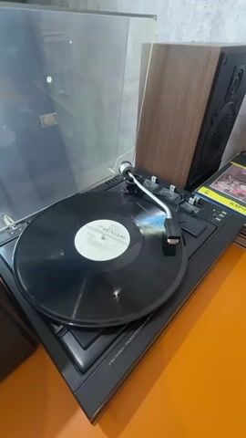 The video displays a close-up shot of a record player. The record player is black and has a vinyl record with a white label playing on it. A tonearm with a needle is positioned on the record. To the right of the record player is a wooden speaker and to the left is a glass cover, likely for the record player. The background appears to be a wall with a gray texture and a small portion of an orange surface is visible at the bottom of the frame. The lighting suggests an indoor environment.