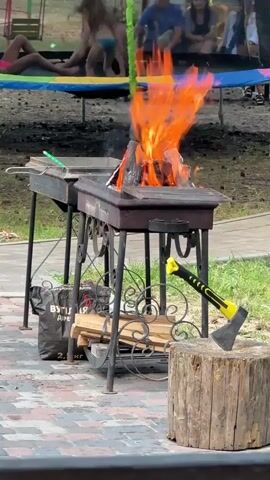A barbecue grill with a fire burning in it sits on a paved surface. An axe rests against a chopping block to the right of the grill. In the background, several people are visible, some sitting and some standing, with a trampoline and some greenery behind them.