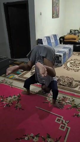 A person indoors is attempting an acrobatic headstand-like move. The person is on hands and head, with legs extended into the air, wearing a gray shirt, gray shorts, and black pants. The person wobbles, loses balance, and falls forward onto their knees. The scene takes place on a pink rug in a room containing sofas, a decorative rug, and a door.