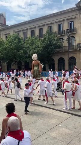 The video shows a square in front of a large, classical-style building with columns and ornate windows. A group of people, most wearing white shirts and red sashes or bandanas, are gathered in the square. Several people are walking, while others appear to be observing. Dominating the scene are two large, elaborate giant figures, known as Gigantes, which are traditional in some European festivals. One Gigante, a female figure, is wearing a patterned green dress and a large, intricate headdress. The other Gigante, a male figure, is dressed in a red and white outfit with a regal cape and a tall, conical hat. The Gigantes are being maneuvered through the crowd, presumably by people hidden within them. The atmosphere appears festive, with many people holding up phones to capture the spectacle. The ground is paved with stone, and trees are visible in the background. The sky is partly cloudy.