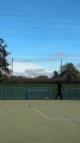 The video shows a person playing soccer outdoors. The person is wearing black clothing and is standing on a light green artificial turf surface marked with white lines, presumably outlining a small soccer field. A blue metal fence surrounds the field, with residential houses and greenery visible in the background. A cloudy blue sky is overhead, with power lines spanning across the frame. The person kicks a soccer ball, sending it out of the frame.