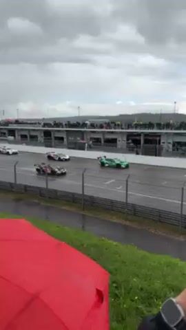 From an elevated perspective, a video captures a race occurring on a wet track. The scene is dominated by a grey, overcast sky. A line of race cars, varying in color and design, speeds along the track, leaving visible spray in their wake. A crowd of spectators is seen standing on a platform above the pit lane, many using umbrellas for protection from the rain. In the foreground, the edge of a red umbrella is visible, along with some grassy terrain. The setting appears to be a professional racing circuit.