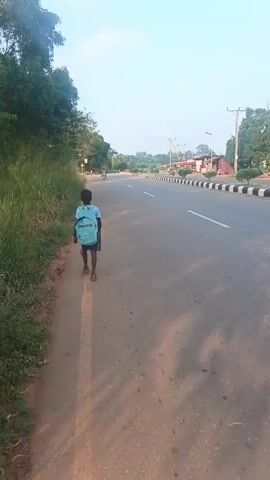 A young boy with a light blue backpack walks on a narrow sidewalk beside a paved road. The road has a white dividing line and black and white striped guardrail.  A motorcycle passes the boy on the road.  Trees and tall grass line the sidewalk to the left.  The sky is pale blue. Buildings are visible in the distance on the right.